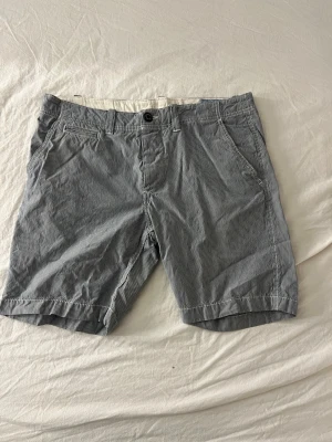 Randiga shorts från Jack & Jones - Säljer ett par randiga shorts från Jack & Jones i blått och vitt. Klassisk modell med fickor fram och bak, bälteshällor och knappstängning. Perfekta för sommaren och tillverkade i mjuk bomull för skön känsla. Står ingen storlek men skulle säga M/L