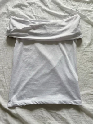 Vit axelbandslös tubtopp - Säljer en enkel och stilren vit tubtopp i mjukt bomullsmaterial. Toppen är axelbandslös och har en bred kant upptill som ger en snygg passform. Perfekt att matcha med jeans eller kjol för en clean look.