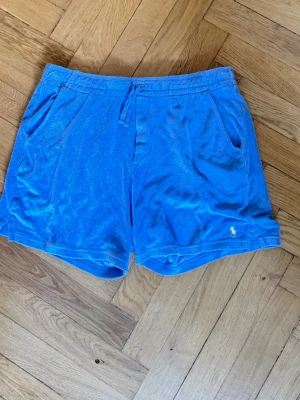 Blå frottéshorts från Ralph Lauren - Säljer ett par blå shorts från Ralph Lauren i mjuk frotté och två sidofickor. Klassisk broderad logga i vitt på ena benet. Perfekta för sommardagar eller att chilla i hemma. Säljer då jag köpte fel storlek Bara att höra av sig vid funderingar! 