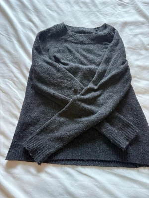 Svart/Mörk grå stickad tröja - En enkel och stilren mörkgrå stickad tröja med rund halsringning och långa ärmar. Tröjan har ribbstickade muddar vid ärmslut och nederkant. Perfekt för lager på lager och passar till det mesta i garderoben.