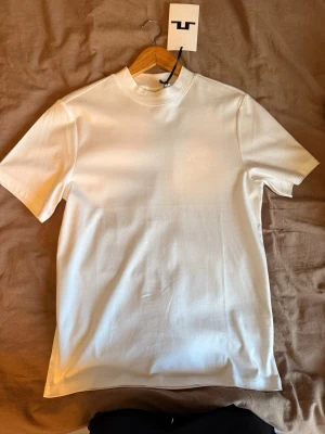 Vit t-shirt från J.Lindeberg - Aldrig använd och med prislapp kvar. Köpt på season i Jönköping. Klassisk vit t-shirt från J.Lindeberg med diskret logga på bröstet. T-shirten har rund hals, korta ärmar och är gjord i mjuk bomull. Perfekt basplagg som passar till allt och har en clean, stilren look. Pris kan diskuteras vid snabb affär! 