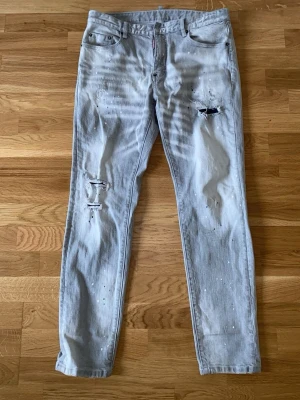 Ljusa slitna jeans från Dsquared2 - Säljer ett par ljusgrå jeans från Dsquared2 med skinny fit och coola slitningar samt färgstänk. Jeansen har klassisk femficksmodell och låg midja. Perfekta för dig som gillar en trendig och avslappnad streetstil.