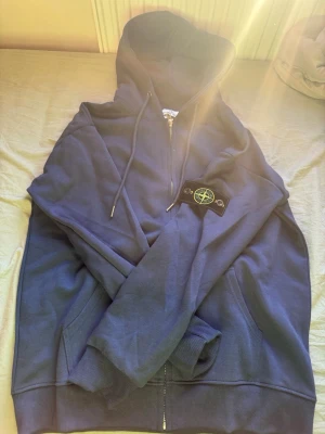Blå jacka dragkedja  från Stone Island - Säljer en jacka från Stone Island med dragkedja och klassisk patch på vänster ärm. Hoodien har huva med dragsko och ribbade muddar vid ärmslut och nederkant. Perfekt för dig som gillar streetwear och vill ha något stilrent och bekvämt. 170cm 55-65kgs M