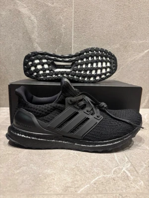 Svarta Adidas Ultraboost sneakers - Säljer ett par helsvarta Adidas Ultraboost sneakers med stickad ovandel och klassiska tre ränder på sidan. Skorna har en bekväm och responsiv Boost-sula samt svart yttersula. Perfekta för dig som gillar stilrena och sportiga sneakers.