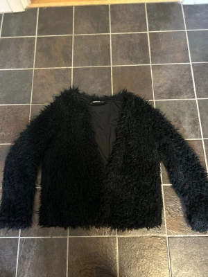 Svart teddyjacka med lång ärm - Säljer en svart teddyjacka med fluffig och mjuk yta. Jackan har lång ärm, rak passform och är helsvart utan synliga detaljer som knappar eller dragkedja. Perfekt för dig som vill ha en mysig och trendig look.