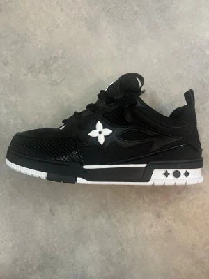 Louis Vuitton svartvit sneaker - Snygga Louis Vuitton sneakers i svart och vitt med ikonisk blomdetalj på sidan. Skon har en låg siluett, meshpaneler och detaljer i läder samt en robust sula med stjärnmönster. Perfekt för dig som vill sticka ut med exklusiv streetstyle.