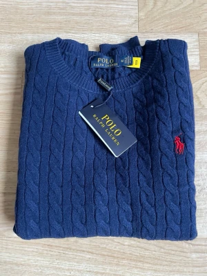 Mörkblå kabelstickad tröja Polo Ralph Lauren - Mörkblå kabelstickad tröja från Polo Ralph Lauren med rund halsringning och klassisk röd logga broderad på bröstet. Tröjan har långa ärmar och ribbade muddar. Perfekt för en stilren och tidlös look.
