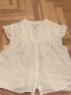 Vit kortärmad blus med knappar från HM - Söt vit blus med korta ärmar och knäppning framtill. Blusen har diskreta broderade prickar och dekorativa veck längs bröstet. Luftigt och lätt material, perfekt för varma dagar. Avslappnad passform och v-ringad hals.