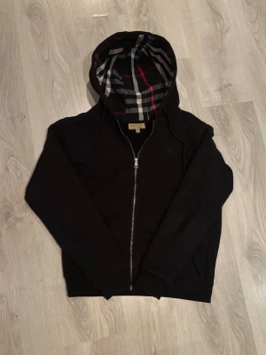 Svart hoodie från Burberry med dragkedja M - Säljer en svart hoodie från Burberry med dragkedja framtill och klassiskt rutigt foder i huvan i svart, vitt och rött. Hoodien har justerbar dragsko i huvan och ribbade muddar vid ärmslut och nederkant. Perfekt för dig som gillar stilrena detaljer och premiumkänsla. Helt perfekt förutom ena snöret som inte har den där lilla plutten. Storlek M men skulle kunna passa S. Kontakta vid frågor om pris 😊
