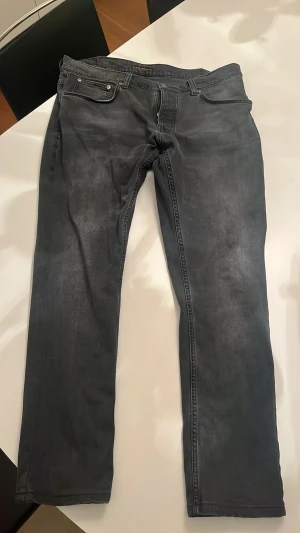 Mörkgrå jeans från Nudie Jeans - Mörkgrå Nudie jeans. Jeansen har diskreta slitningar och Nudie Jeans signatursöm på bakfickorna. Tillverkade i mjuk denim som ger en avslappnad look. Jeansen är upplagda så att de är lite smalare i midjan och lite kortare så storleken är egentligen runt 34/31