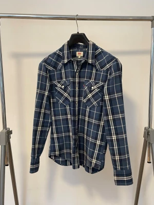 Blå rutig skjorta från Levi's - Väldigt snygg blå rutig skjorta från Levi's med vita och grå rutor. Skjortan har två bröstfickor med knappar och klassisk krage. Perfekt för en avslappnad och cool stil.