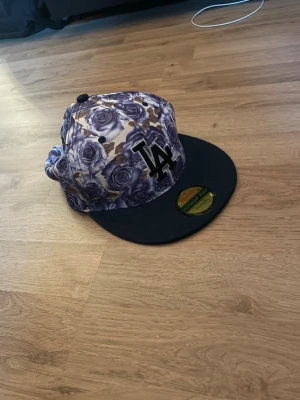 LA snapback med lila rosor - Säljer en snapback-keps med LA-logga framtill och ett unikt mönster av lila rosor över hela kepsen. Skärmen är svart och modellen är justerbar baktill. Perfekt för dig som vill sticka ut med en färgstark och trendig accessoar.