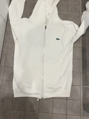 Vit hoodie med dragkedja från Lacoste - Vit hoodie med dragkedja från Lacoste, klassisk krokodillogga på bröstet. Mjuk bomullskänsla, huva med dragsko och två fickor framtill. Perfekt för en avslappnad och stilren look.