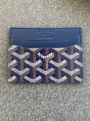 Goy4rd plånbok - Snygg blå korthållare med mönster i vitt och brunt. Tillverkad i läder med flera fack för kort. Perfekt accessoar för dig som vill ha något både stilrent och exklusivt i fickan.