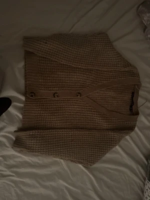 Beige stickad kofta med knappar - Mysig beige stickad kofta med v-ringning och stora knappar framtill. Koftan har ett grovt, rutigt stickmönster och långa ärmar. Perfekt att slänga över en t-shirt eller topp för en avslappnad look.