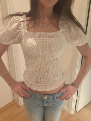 Vit smockad blus med volang - Supersöt vit blus med smockad kropp, puffiga korta ärmar och volangdetaljer vid halsringning och nederkant. Blusen har fyrkantig urringning och är lätt croppad, perfekt till jeans eller kjol.