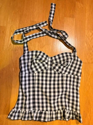 Svartvit rutig korsett-topp med volang - Säljer en snygg korsett-topp med svartvitt rutigt mönster och knytband som går runt halsen. Toppen har en söt volangkant längst ner och smickrande formade kupor framtill. Perfekt för dig som gillar trendiga och unika plagg!