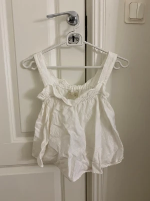 Vit volanglinne med breda axelband - Säljer ett vitt linne med breda axelband och volangdetaljer. Toppen har en luftig och lös passform med rynkad kant upptill. Perfekt för sommardagar och enkel att styla med jeans eller kjol. Materialet känns lätt och svalt.