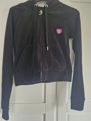Svart Juicy Couture hoodie med hjärta - Cool svart hoodie från Juicy Couture med glittrande rosa hjärta på bröstet och stort Juicy Couture-tryck med krona och hjärta på ryggen. Mjuk velour, dragkedja, huva med snören och stora fickor framtill. Perfekt för dig som gillar glam och comfy vibes. Saknas ena metallpluppen på snöret