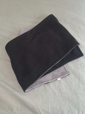 Svart och grå fleecehalsduk - Mjuk och varm halsduk i fleece med ena sidan svart och andra sidan grå. Rektangulär form och enkel design som passar till de flesta ytterkläder. Perfekt för kalla dagar när du vill hålla dig varm och bekväm.