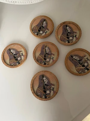Sex runda träunderlägg med konstmotiv - Säljer ett set med sex runda underlägg i trä, dekorerade med färgstarka motiv av kvinnor i traditionella kläder. Underläggen har en varm brun ton och detaljerade illustrationer i lila, svart och beige. Perfekta för att skydda bordet och ge en unik touch till hemmet.Set med 6 underlägg i trä med indiska motiv. Handgjord. Det är är gjort av trä och glasskiva på . Det finns en som är glasset är sprucken i mitten och en som är trä liten sprucken . 