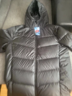 Svart dunjacka från Helly Hansen - Snygg svart dunjacka från Helly Hansen med huva och dragkedja framtill. Jackan har breda, horisontella kanaler och en stilren design som håller dig varm under kalla dagar. Perfekt för dig som vill ha en klassisk och funktionell vinterjacka.