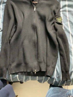 Svart ziptröja från Stone Island - Svart stickad ziptröja från Stone Island med ribbade muddar och klassisk logotyp-patch på vänster ärm. Tröjan har hel dragkedja framtill och är tillverkad i ett grovstickat material som ger en cool och stilren look.