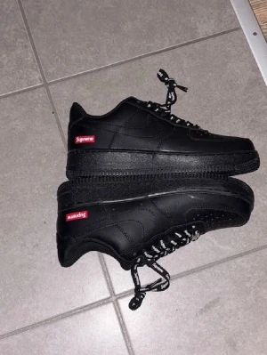 Svarta Supreme x Nike Air Force 1 - Säljer ett par svarta Supreme x Nike Air Force 1 sneakers i läder. Skorna har Supreme-logga i rött på sidan och svarta skosnören med vit Supreme-text. Klassisk Air Force 1-design med perforerad tå och låg profil.