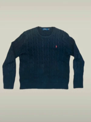 Polo Ralph Lauren - Kabelstickad mörkblå tröja från Polo Ralph Lauren med rund halsringning och röd broderad logga på bröstet. Tröjan har ribbade muddar vid ärmslut och nederkant. Klassisk och tidlös design som passar till mycket.