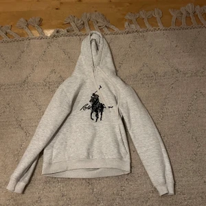 Grå hoodie från Ralph Lauren - Snygg ljusgrå hoodie från Ralph Lauren med det klassiska stora polospelar-trycket i svart på bröstet. Hoodien har huva med vita dragsnören och en känguruficka framtill. Perfekt för en avslappnad och trendig stil. Jättebra skick nästan aldrig använd. Storlek M men passar även S