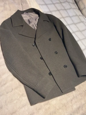 Dressman peacoat - Stilren mörkgrön skepparkavaj med dubbelknäppning och breda slag. Jackan är tillverkad i ett tjockt ullmaterial och har en klassisk, rak passform. Perfekt för dig som vill ha en tidlös look med lite edge. Helt ny, Nypris 2000kr från dressman storlek 48 M. 