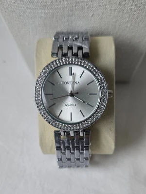 Silverfärgad klocka från Contena - Stilren klocka från Contena i silver med glittrig ram runt urtavlan. Klockan har en klassisk rund form, länkarmband i metall och minimalistisk urtavla utan siffror. Perfekt accessoar för dig som gillar en elegant look.