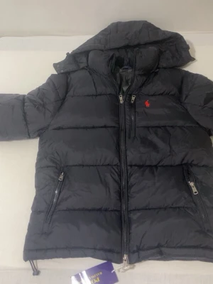 Svart pufferjacka från Polo Ralph Lauren - Snygg svart pufferjacka från Polo Ralph Lauren med huva och dragkedja framtill. Jackan har två sidofickor med dragkedja och en bröstficka. Klassisk röd logga broderad på bröstet. Perfekt för kalla vinterdagar och riktigt skön att bära. Den är helt ny har aldrig använts.