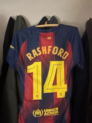 FC Barcelona Rashford matchtröja - Säljer en FC Barcelona matchtröja från Nike med Rashford och nummer 14 på ryggen. Tröjan är blå och röd med gula detaljer, klubbmärke och sponsorloggor på bröstet. Tillverkad i lätt och ventilerande material, perfekt för fotbollsträning eller support.