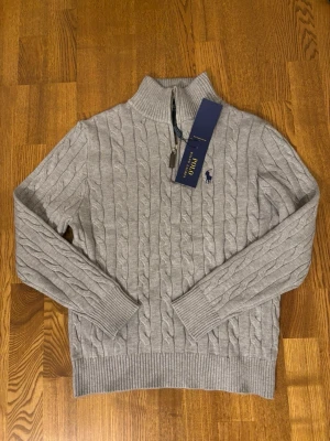 Grå kabelstickad tröja Polo Ralph Lauren - Snygg grå kabelstickad tröja från Polo Ralph Lauren med halv dragkedja och hög krage. Tröjan har ribbade muddar och nederkant samt den klassiska mörkblå loggan broderad på bröstet. Perfekt för dig som gillar stilrena och tidlösa plagg.