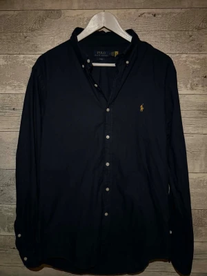 Mörkblå skjorta från Polo Ralph Lauren - Klassisk mörkblå skjorta från Polo Ralph Lauren med button-down krage och gul broderad logga på bröstet. Skjortan har långa ärmar, vita knappar och är tillverkad i mjuk bomull. Perfekt för en clean och stilren look.