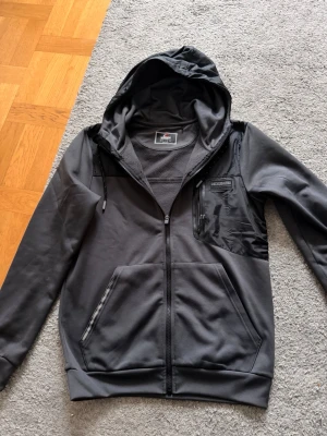 Mörka grå jacka från MCKENZIE - Mörkgrå jacka från MCKENZIE med huva och dragkedja framtill. Jackan har en bröstficka med dragkedja och två sidofickor. Tillverkad i mjukt syntetmaterial och har långa ärmar med ribbade muddar. Perfekt för dig som gillar en sportig och clean stil.