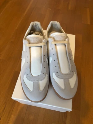 Maison Margiela Replica sneakers vita - Maison Margiela Replica sneakers i vitt och ljusgrått med klassisk design. Skorna har en kombination av läder och mocka, gummisula och ikonisk Margiela-tunga med präglad logotyp. Perfekt för dig som gillar stilrena och trendiga sneakers. Köpt från farfetch, snören är upplösta.