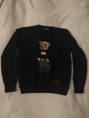 Mörkblå Polo Ralph Lauren björntröja - Stickad mörkblå tröja från Polo Ralph Lauren med ikonisk björn på bröstet, klädd i jeans och tröja med amerikansk flagga. RL-broderi i rött nertill. Rund hals och ribbade muddar. Perfekt för dig som gillar klassisk preppy-stil. Bröst:45cm längd:56cm arm:56cm