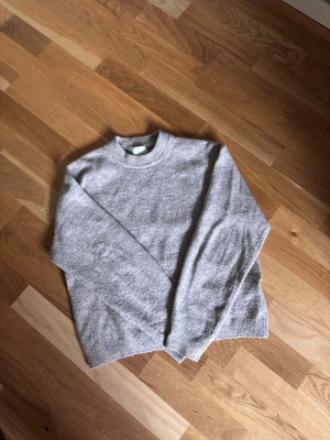 Ljusgrå stickad tröja i ullmix - Mysig ljusgrå stickad tröja med rund hals och ribbade muddar vid ärmslut och nederkant. Tröjan har en avslappnad passform och är tillverkad i en mjuk ullblandning som passar perfekt för kyligare dagar.