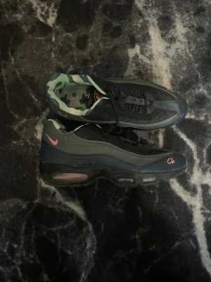 Nike Air Max 95 x Cx svarta sneakers - Nike Air Max 95 x Cx sneakers i svart och mörkgrått med rosa detaljer. Skorna har en unik camo-mönstrad insida och synlig Air Max-dämpning i sulan. Klassisk vågig design och meshpaneler för extra ventilation. Perfekta för dig som vill sticka ut. PRISET KAN DISKUTERAS!!! PÅ GRUND AV LITE SKADOR!