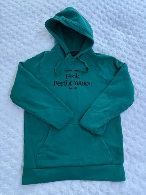 Grön hoodie från Peak Performance - Snygg grön hoodie från Peak Performance med broderad logga på bröstet och känguruficka framtill. Klassisk huva med dragsko och mjukt bomullsmaterial. Perfekt för en chill och sportig stil. Har väldigt lätta slitningar vid fickan men knappt synliga.
