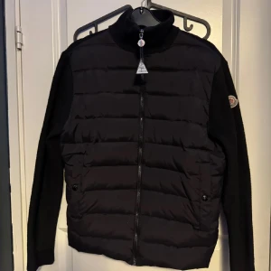Svart dunjacka från Moncler - Säljer en snygg svart dunjacka från Moncler med långa ärmar och dragkedja framtill. Jackan har Moncler-logga på ärmen och röda detaljer längs sidorna. Qr och tagen funkar! Skriv vid minsta fundering🍾 pris är ej hugget i sten