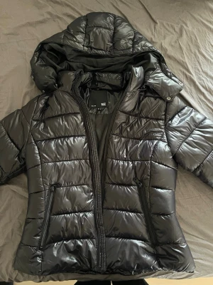 Svart pufferjacka från 157 - Snygg svart populär pufferjacka från 157 med avtagbar huva och två dragkedjeförsedda fickor framtill. Jackan har quiltad design och är tillverkad i glansigt syntetmaterial som ger en modern look. Perfekt för kalla dagar när du vill hålla dig varm och stilren. 