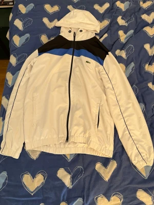 Vit vindjacka från Lacoste med huva - Snygg vit vindjacka från Lacoste med svart och blå detalj upptill. Jackan har huva, dragkedja framtill och två fickor. Klassisk krokodil-logga på bröstet och elastiska muddar. Perfekt för blåsiga dagar och sportig stil. Passar L också 