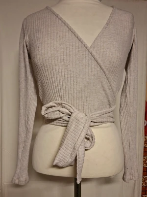 Beige omlottopp med knytband - Trendig beige omlottopp i ribbat material med långa ärmar och breda knytband i midjan. Djup v-ringning framtill och figurnära passform. Perfekt för lager på lager och enkel att styla till olika looks.