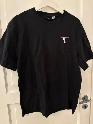 Svart Snoopy t-shirt Beach Beagle - Svart t-shirt med tryck av Snoopy och texten 'Beach Beagle' på ryggen. Framtill finns en liten Snoopy med rosa surfplatta. T-shirten har rund hals och korta ärmar, tillverkad i mjuk bomull. Perfekt för dig som gillar retro och surfvibbar.