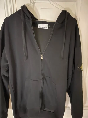 Svart zip hoodie från Stone Island - Säljer en svart hoodie från Stone Island med dragkedja framtill, stor huva och känguruficka. Klassisk logopatch på vänster ärm. Tillverkad i mjuk bomull för skön känsla och avslappnad stil. Perfekt för dig som gillar streetwear och vill ha något stilrent.