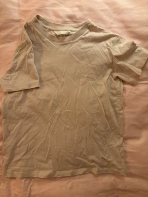 Beige basic t-shirt från H&M - En enkel och stilren beige t-shirt från H&M i mjuk bomull. T-shirten har rund halsringning och korta ärmar, perfekt för en avslappnad och clean look. Passar till allt och är ett måste i garderoben.