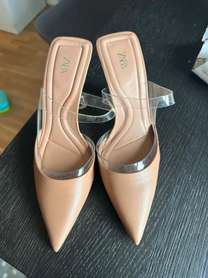 Beige pumps med transparent rem Zara - Snygga beige pumps från Zara med spetsig tå och transparenta plastremmar. Skorna har en stilren design och öppen häl, vilket ger en modern look. Tillverkade i konstläder och plast, perfekta för dig som gillar trendiga detaljer.